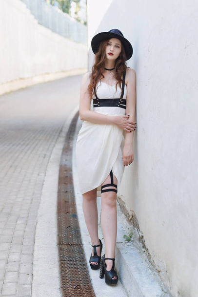 Fiatal gyönyörű szexi nő visel divatos ruhát, fehér ruha, fekete kalap és bőr kardöv. Hosszú hajú barna pózol a város utcájában egy napsütéses napon. Kültéri divat fotózás. - Fotó, kép