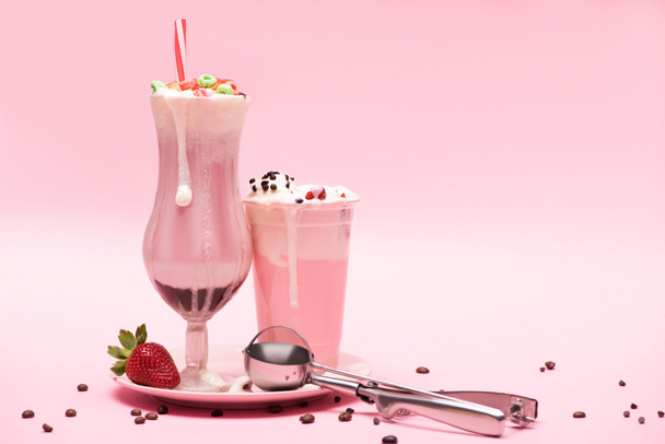 Copo descartável e vidro de milkshakes, morango e colher em prato com grãos de café em rosa
  - Foto, Imagem