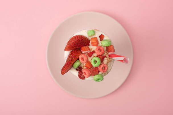 Top view of milkshake with drinking tube, strawberry halves and candies on plate on pink background - Photo, Image