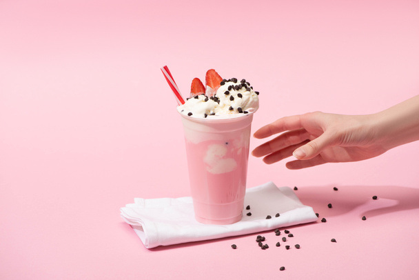 Częściowy widok kobiecej ręki z jednorazowym kubkiem shake 'a z czekoladowymi kąskami i truskawką na serwetkach na różowo - Zdjęcie, obraz