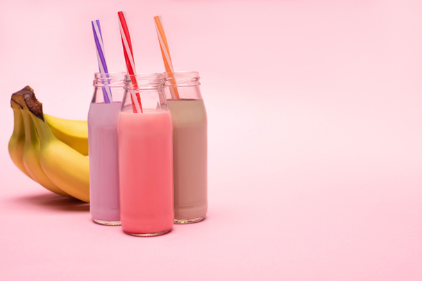Flessen bessen, aardbeien en chocolade milkshakes met drinkrietjes bij bananen op roze achtergrond - Foto, afbeelding