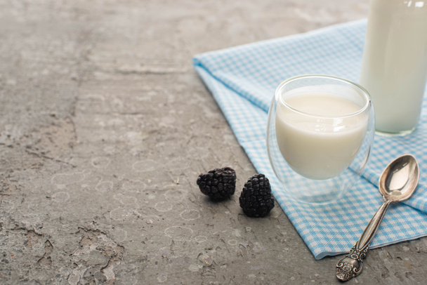 Glass and bottle of homemade yogurt with teaspoon on cloth near blackberries on grey background - Foto, afbeelding