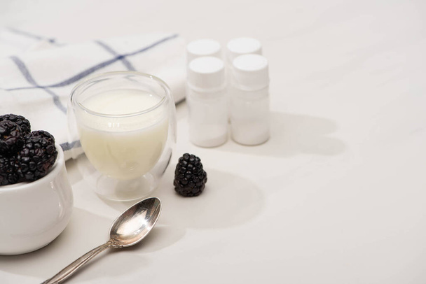 Vista de alto ângulo de colher de chá, copo de iogurte e tigela de açúcar com amoras perto de recipientes com culturas iniciais em branco
 - Foto, Imagem
