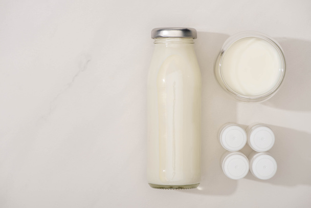 Vista dall'alto di bottiglia e bicchiere di yogurt fatto in casa vicino a contenitori con colture di antipasto su sfondo bianco
 - Foto, immagini