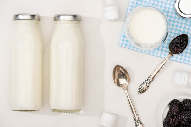 Vista dall'alto di bottiglie e bicchiere di yogurt su stoffa, cucchiaini, more e contenitori con colture di antipasto su bianco
 - Foto, immagini