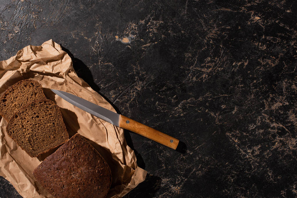 vrchní pohled na čerstvě nakrájený bochník hnědého chleba s nožem na papíře na kamenném černém povrchu - Fotografie, Obrázek