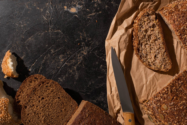 top view of cut whole grain bread on paper near knife on stone black surface - Fotó, kép