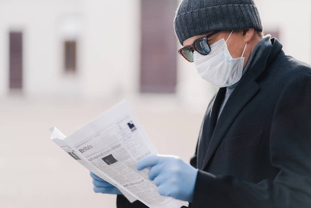 Карантин для коронавируса. Горизонтальный снимок взрослого мужчины носит защитную маску и одноразовые перчатки, читает свежую газету, узнаёт новости о пандемической ситуации. Концепция профилактики инфекции
 - Фото, изображение
