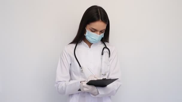 Φιλική γυναίκα γιατρός σε λευκό ιατρικό παλτό γάντια και προστατευτική μάσκα, γράφει τη διάγνωση στην κάρτα πελατών, - Πλάνα, βίντεο