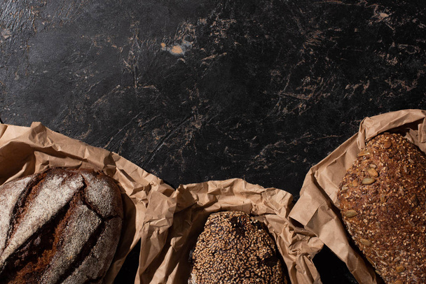 top view of fresh baked whole grain bread loaves in paper on stone black surface - Photo, Image