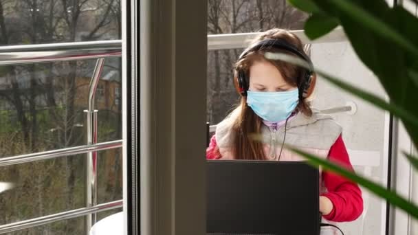 teenager girl in mask and headphones, typing, working on laptop, on open balcony. spring sunny day. distance learning during quarantine. stay, study at home. coronavirus epidemic. - Imágenes, Vídeo