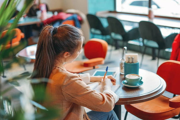 Young brunette girl student doing homework in notebook with coffee at cafe - Photo, Image