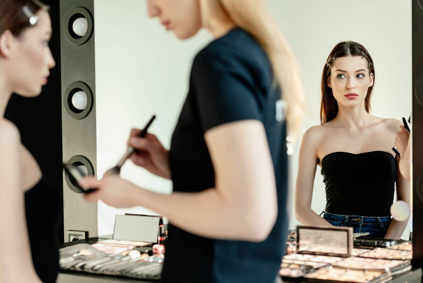 επιλεκτική εστίαση του όμορφου μοντέλου κοιτάζοντας καθρέφτη κοντά καλλιτέχνη μακιγιάζ  - Φωτογραφία, εικόνα