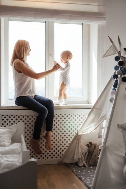 Retrato de una joven madre linda y sonriente y su hijo jugando cerca de la ventana en una habitación blanca brillante. Día soleado afuera. Amor de madre verdadera
 - Foto, imagen