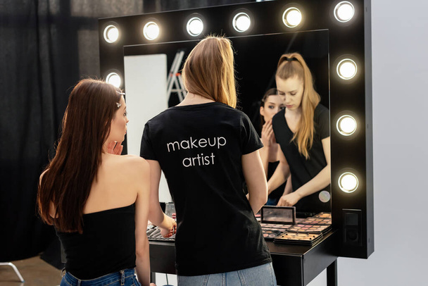 woman in black t-shirt with makeup artist lettering standing near model and decorative cosmetics  - Фото, изображение