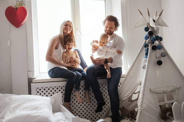 Happy smiling modern caucasian family playing and spending time in a bright white room near the window. Sunny day. Perfect parenting relationship. Parents of two cute boys. - Foto, afbeelding