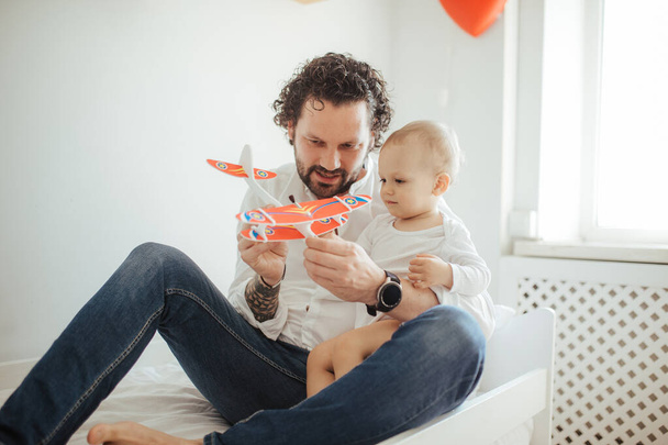Young father o a son playing with a toy plane. Happy family, two boys in a bright sunny room spending time together on a bed. - Photo, Image