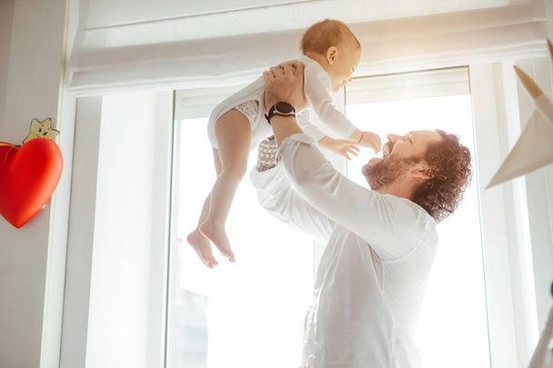 Father playing with cute newborn baby boy near the window in a sunny bright room. Loving father rising his son up and makes him smile. Enjoying sunny days at home. Perfect parenting relationship - Photo, Image
