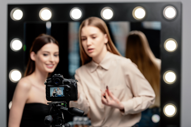 foco seletivo da câmera digital com o artista de maquiagem segurando o gloss perto do modelo positivo na tela
  - Foto, Imagem