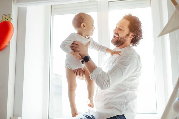 Father playing with cute newborn baby boy near the window in a sunny bright room. Loving father rising his son up and makes him smile. Enjoying sunny days at home. Perfect parenting relationship - Foto, immagini