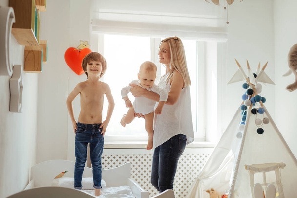 Young blonde mother of two boys playing in a bright room. Sunny day outside. Caucasian family spending time together. Older brother jumping on the bed. Little brother in mother hands. Friendly family - Fotó, kép
