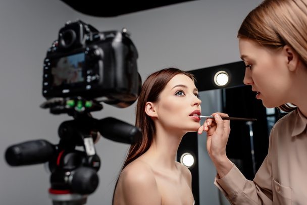 selective focus of makeup artist applying lip gloss on lips of attractive model near digital camera on grey  - Photo, Image