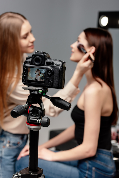 selective focus of tripod with digital camera near happy makeup artist applying face powder on beautiful model  - Fotografie, Obrázek