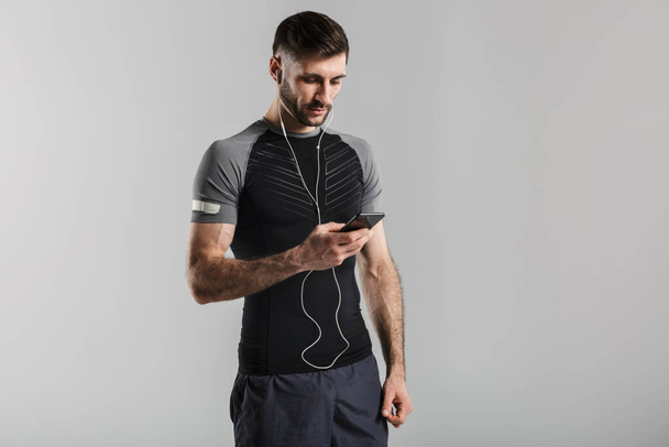 Image of a handsome strong sportsman isolated over grey wall background using mobile phone listening music with earphones. - Foto, immagini
