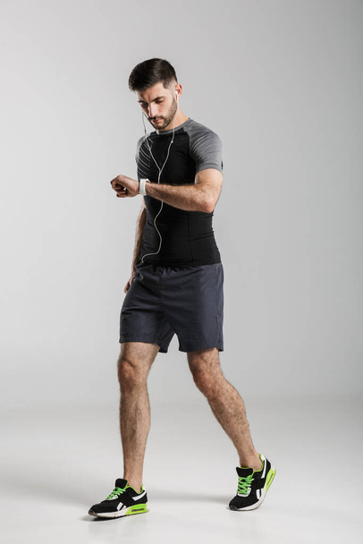 Image of young unshaven sportsman using smartwatch and earphones isolated over gray background - Photo, image