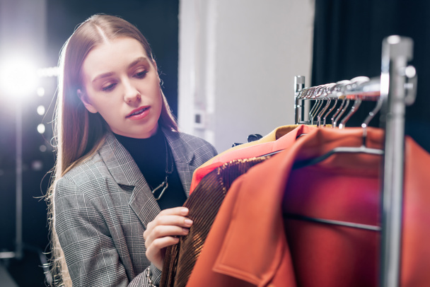 selective focus of attractive stylist looking at fashionable clothing on hangers  - 写真・画像