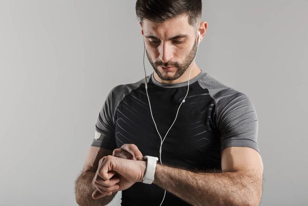 Image of young unshaven sportsman using smartwatch and earphones isolated over gray background - Фото, изображение