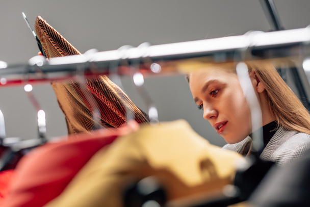 selective focus of beautiful stylist looking at fashionable clothing on hangers  - Valokuva, kuva