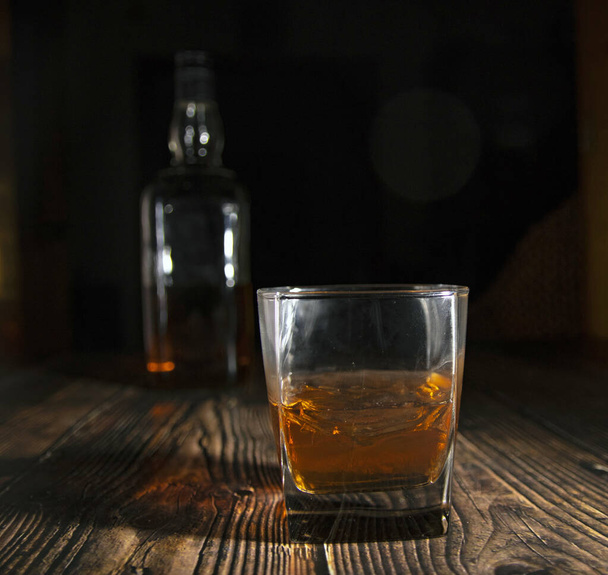 Стакан виски с кубиками льда, бутылка на деревянном столе. Старый стол со светом и бокалом крепкого напитка
. - Фото, изображение