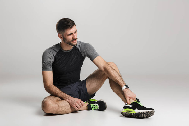 Image of unhappy athletic sportsman with injury sitting while working out isolated over gray background - Fotó, kép