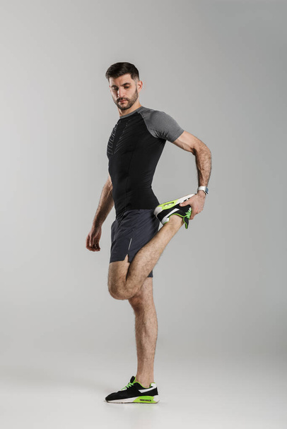 Image of young athletic man in sportswear stretching her leg while working out isolated over gray background - Foto, afbeelding