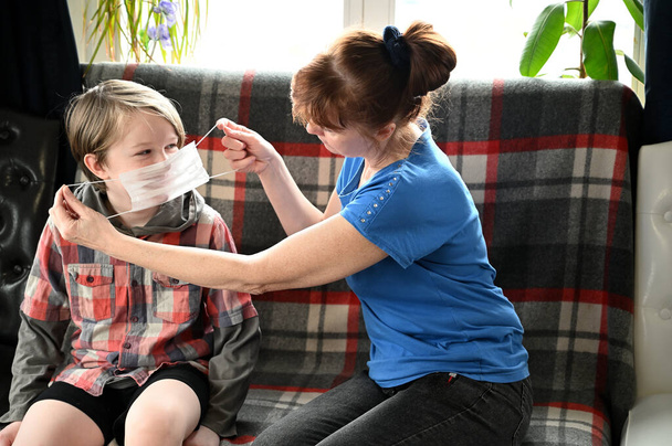 Photo of mom and boy sitting on the sofa in the home interior. Mother teaches quarantine her son to wear a medical mask - Foto, Imagen