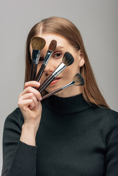 young makeup artist covering face with cosmetic brushes isolated on grey  - Foto, Imagem