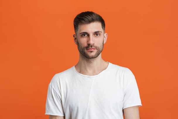 Image of young bearded man wearing basic white t-shirt looking at camera isolated over orange background - Foto, Imagem