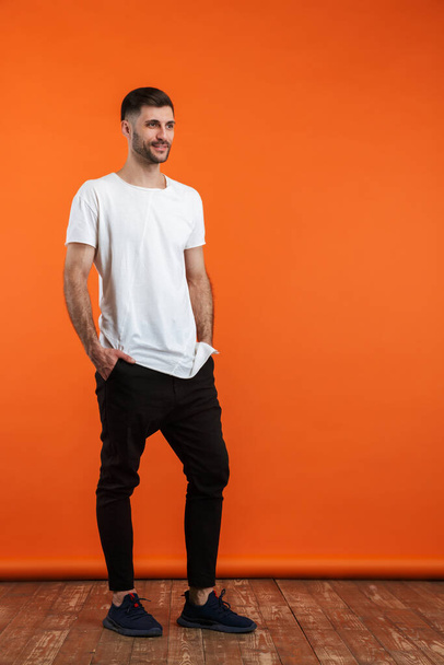 Image of young bearded man wearing basic white t-shirt smiling and looking aside isolated over orange background - Foto, imagen