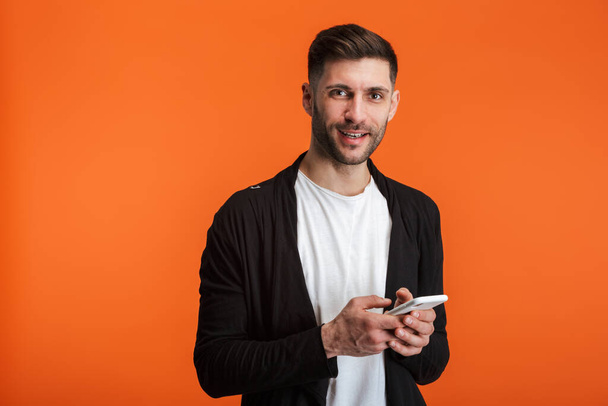 Image of young bearded man wearing basic clothes smiling and holding cellphone isolated over orange background - Photo, Image