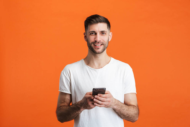 基本的な服を着て笑顔とオレンジの背景に隔離された携帯電話を保持している若い髭の男のイメージ - 写真・画像