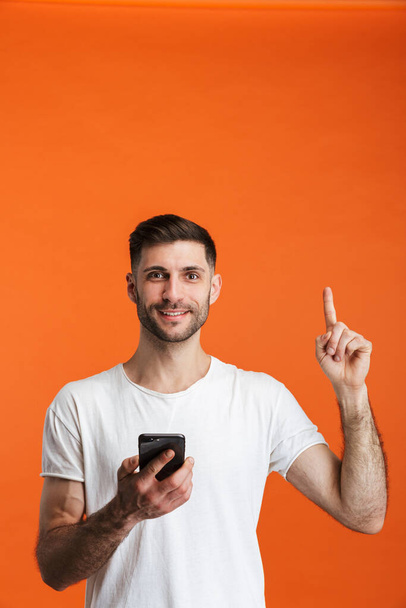 Image of young bearded man wearing basic clothes holding cellphone and pointing finger upward isolated over orange background - Photo, Image