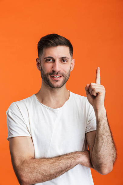 Image of young bearded man wearing basic clothes smiling and pointing finger upward isolated over orange background - Foto, Bild