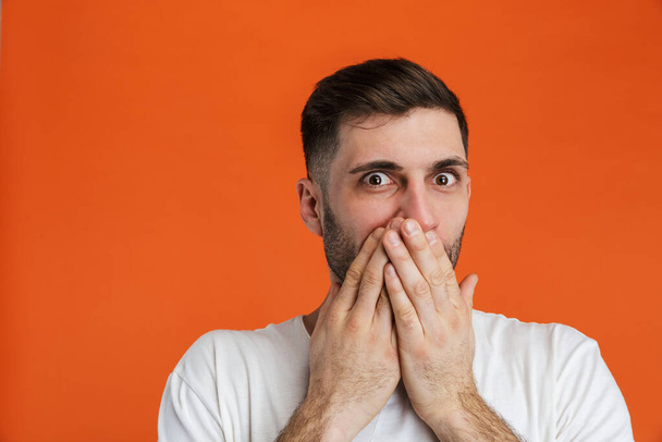 Image of young terrified man wearing basic white t-shirt covering his mouth with hand isolated over orange background - Foto, immagini