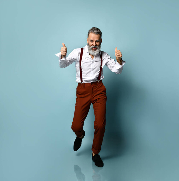 Aged male in white shirt, brown pants and suspenders, black loafers. He is showing thumbs up, walking on blue studio background - Photo, image
