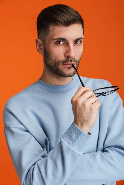 Image of young bearded handsome man wearing sweater holding his eyeglasses isolated over orange background - Foto, immagini