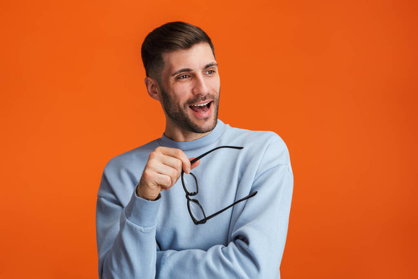 Image of young bearded handsome man wearing sweater holding his eyeglasses isolated over orange background - Zdjęcie, obraz