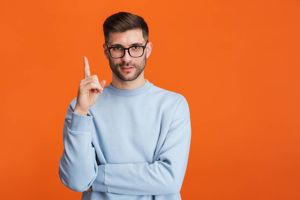 Image of young bearded handsome man wearing eyeglasses smiling and pointing finger upward isolated over orange background - Foto, Imagem
