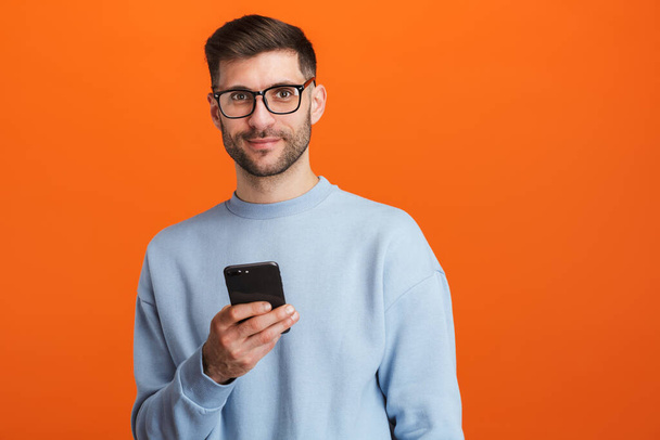 Image of young bearded handsome man wearing eyeglasses holding cellphone isolated over orange background - Φωτογραφία, εικόνα