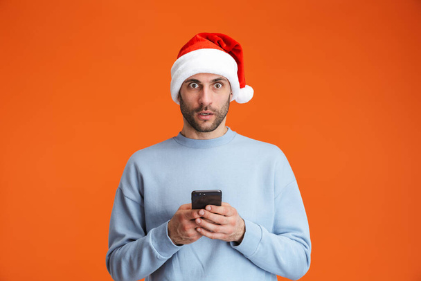 Image of a shocked young man in christmas santa hat posing isolated over orange wall background holding mobile phone. - Photo, image